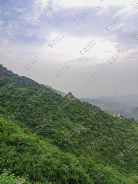 A natural scene capturing a mountain covered with dense green trees under a cloudy blue sky. In the middle of the image, there is a stone structure on top of the mountain, with other tree-covered hills visible in the background.