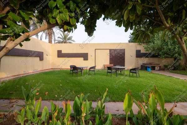 An image of an outdoor courtyard featuring a green lawn and flowering plants in the foreground. The garden includes two plastic tables surrounded by black chairs. The back wall is beige with brown rectangular decor. Trees surround the area, and there is a blue trash bin in the right corner.