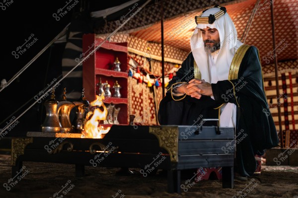 A man sitting in a traditional tent wearing a white and black traditional Arabic robe with a headband. In front of him, there is a lit fire pit and several silver coffee pots. The tent is adorned with traditional Arabic patterns and has shelves with coffee pots and traditional items.