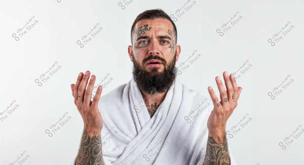 Portrait of a bearded man with tattoos on his face and hands. He is wearing a white robe and appears to be in a contemplative pose. The man is looking forward with his hands slightly open. The background is white and clean.