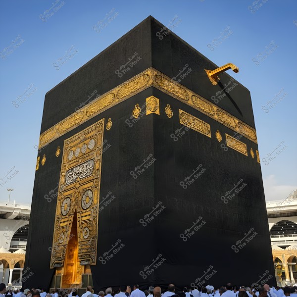 The image shows the Kaaba in Mecca, Saudi Arabia. The Kaaba is covered with a black cloth adorned with golden embroidery. At the bottom of the image, a group of people wearing white ihram garments are visible, indicating the performance of Umrah or Hajj in the holy mosque. The background features a clear blue sky.