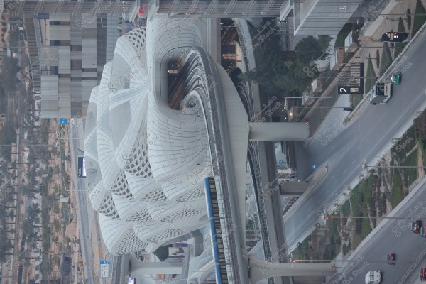 An image of a train station with a modern and innovative architectural design, featuring a white exterior with distinctive geometric patterns. Surrounding roads and bridges intersect near the station, with several small cars driving on the nearby roads. There are many trees and green areas adding vibrancy to the urban scene.