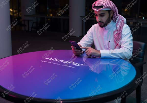Image of a man wearing a white thobe and red headscarf, seated at an illuminated table. The individual is using a mobile phone, and the table displays the word \"AlimaPay\" featuring a gradient of blue to pink. The setting appears modern, suggesting the person might be from a Gulf country due to the traditional attire style.