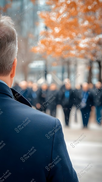 Image of a man photographed from behind wearing a dark blue coat standing under light snowfall. In the background, there is a blurred group of people and trees with orange autumn leaves. The weather appears cold with light snowflakes falling.