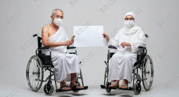 An elderly man and woman are seated in wheelchairs, both wearing white ihram clothing typically worn during the Hajj pilgrimage, along with medical masks. The man on the left is dressed in the traditional ihram attire, while the woman on the right wears a white robe and a white scarf covering her head. They are holding a blank white board between them. The background is white and neutral.