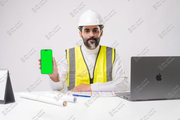 صورة لرجل يجلس على مكتب وهو يرتدي خوذة بيضاء وسترة أمان صفراء فوق الثوب التقليدي. يبدو أنه من خلفية خليجية محتمل أن يكون من السعودية. يمسك الهاتف بشاشة خضراء في يده اليسرى, وعلى المكتب يوجد جهاز كمبيوتر محمول, وأوراق تخطيط, وقلم, ودفتر تقويم.