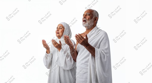 The image shows an elderly man and woman standing in a prayer position. Both are wearing traditional white Ihram clothing. The background is pure white, emphasizing the focus on the individuals. The man is dressed in a white wrap-around garment, while the woman is wearing a white hijab and robe.