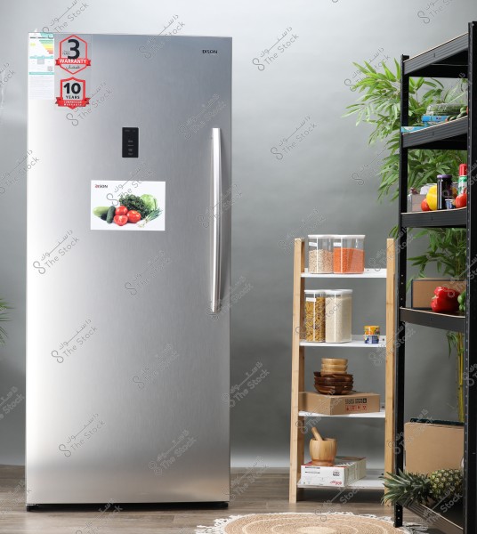The image features a large silver metal refrigerator with a sticker of vegetables in the middle, and warranty stickers indicating 3 and 10 years on the top left. Next to the refrigerator, there is shelving with transparent plastic containers filled with grains and pasta. There are also wooden bowls and cardboard boxes. The adjacent shelf contains various fruits, vegetables, and canned goods.