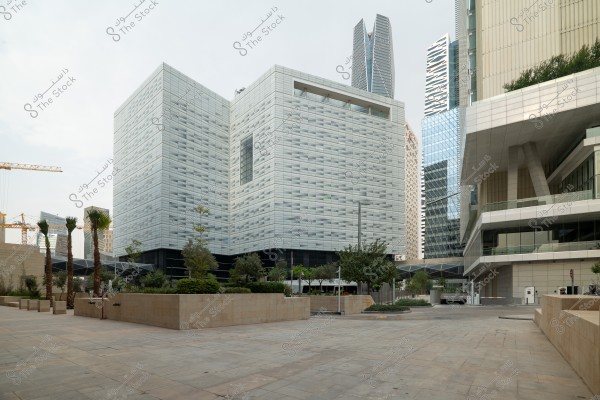 Image of an urban scene in a modern commercial area. A large building with glass facades and intricate architectural design is in the center, surrounded by skyscrapers. In the foreground, there is a small garden planted with trees, alongside a stone pavement. Construction cranes are visible in the background, suggesting ongoing development in the area.