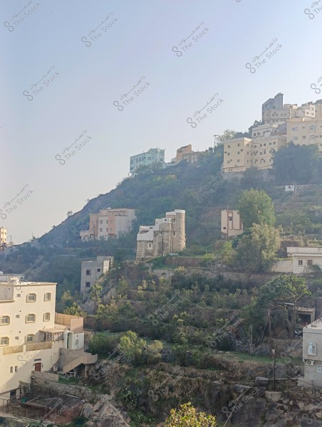 The image depicts a mountainous landscape with several traditional buildings scattered across the mountainside. The buildings are painted in light colors such as beige and light blue, with small stone houses visible among the dense greenery covering the mountain. The sky is clear and blue, creating a serene backdrop for the picturesque scene.