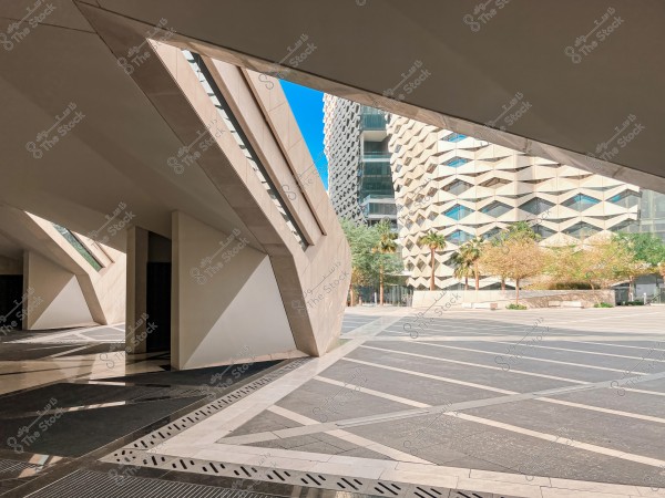A modern architectural view showcasing a building with an innovative design characterized by complex geometric patterns on its exterior. The building is surrounded by a wide open area paved with intersecting shapes. In the background, palm trees and greenery are visible, adding a sense of life and harmony with the contemporary architectural elements.