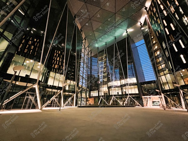 Image of a modern building with distinctive architectural design featuring glass and metal. The building is fully visible in the evening, illuminated from within with warm lights. The ceiling is geometrically designed with innovative glass panels reflecting the blue sky. Tall metal columns add to the contemporary design. Other skyscrapers are visible in the background through the glass.