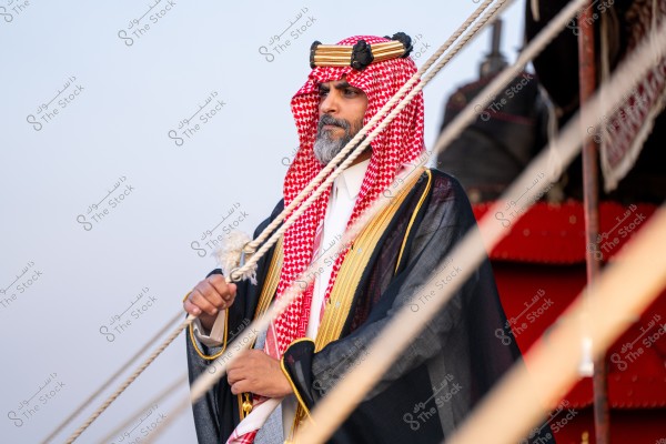 An image of a man wearing traditional Saudi attire, including a white robe, a red and white checked shemagh, and a black bisht, standing beside a set of ropes. He appears to be on the deck of an old ship or boat. The background shows part of the sky and wooden elements.