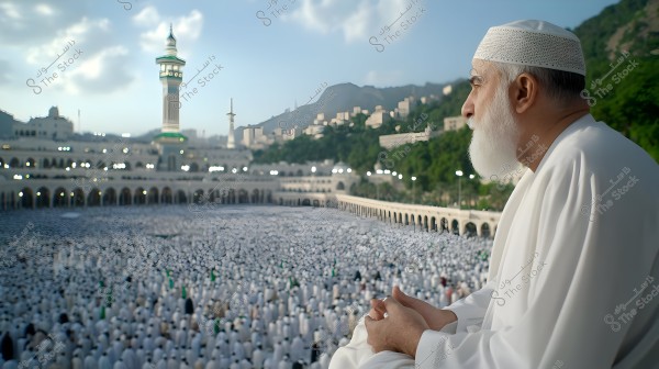 A man wearing a white robe and a white cap is standing beside the Grand Mosque in Mecca. The background shows minarets, buildings, and surrounding mountains with a large crowd of people dressed in white in the expansive courtyard.