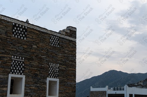 An ancient building built of stones in the middle of a group of windows surrounded by white paint while the sky looks cloudy during sunset, Rijal Almaa Heritage Village in the Asir region