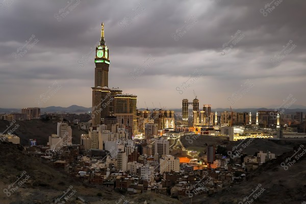 منظر ليلي لمدينة مكة المكرمة, يظهر فيه برج ساعة مكة المشهور بإضاءته الخضراء المميزة, والمباني المحيطة به تحت سماء غائمة.