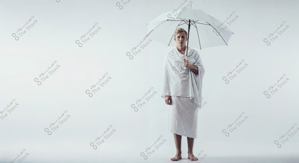 Image of a man standing and holding a white umbrella, wearing a Hajj ihram, which consists of two pieces of white cloth. The background is plain white, and the man is barefoot.