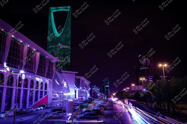A night view of Riyadh, Saudi Arabia, showing the Kingdom Tower illuminated in green in the city center. Surrounding buildings are lit with neon lights in various colors, and cars speed along the road, creating a streaking light effect.