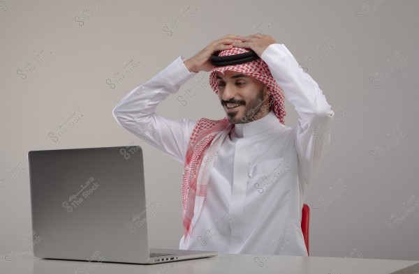 An image of a man sitting in front of a laptop, wearing a white thobe, ghutra, and agal. He appears pleased and holds the agal with both hands. This attire is traditional in the Gulf region.