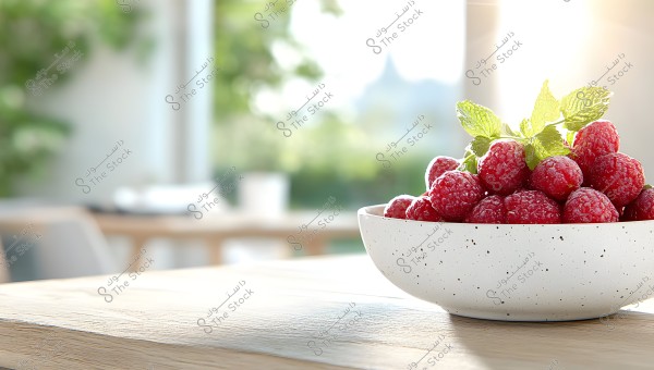 A white bowl filled with fresh red raspberries, topped with green mint leaves, placed on a wooden table surface. The background is blurred with green trees and bright sunlight.
