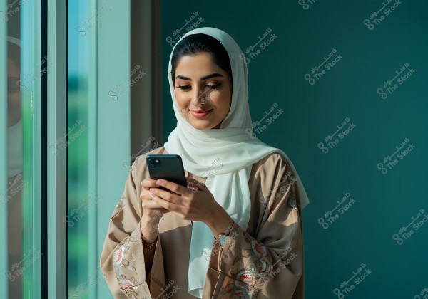 A portrait of a woman wearing a white hijab and a beige abaya with colorful embroidery, standing near a window and using a smartphone. She appears to be smiling, enjoying what she sees on the phone. The background features a blue wall, adding a calm touch to the image.