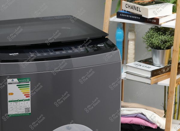 The image shows a modern grey washing machine with a digital control panel on top. To the right of the washing machine, there is a wooden shelf with various covered books, bottles of liquids, and a decorative plant in a pot. There is also a stack of folded clothes on the lower shelf.