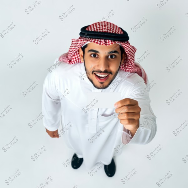 A photo of a man wearing traditional Saudi attire, including a thobe and a red and white checked keffiyeh. The man is pictured from above, holding a blank white card with a surprised expression on his face. The background is white, making the image bright and clear.