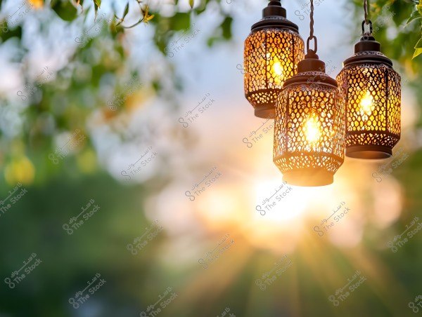 Three decorative metal lanterns illuminated and hanging outdoors from trees, reflecting the sunset light through the green leaves. The scene provides a warm, tranquil glow, with a clear contrast between the intricate details of the lanterns and the blurred background of nature.