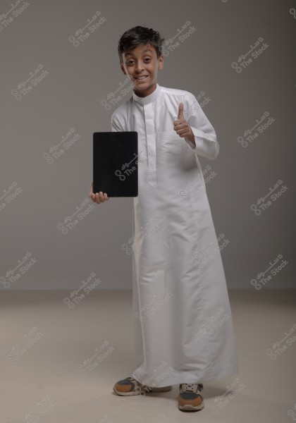 A photo of a boy wearing a traditional white thobe, smiling and giving a thumbs-up. He is holding a tablet in his left hand. He is wearing sneakers and the background is a simple gray. The boy appears to be from the Middle East.