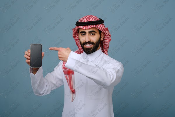 بورتريه شاب سعودي يرتدي الزي السعودي يشير إلى هاتفه ويتفاعل معه، وتظهر على ملامحه الدهشة ، على خلفية سماويه