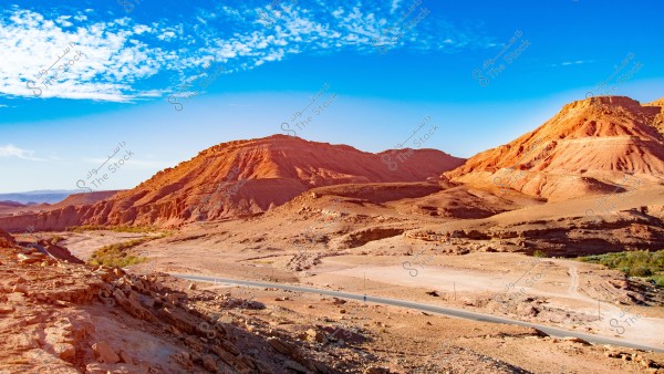 A natural landscape of rocky mountains with red hues in an open desert environment. A road runs through the area below, with a clear blue sky and light clouds in the background. The rugged mountainous terrain and geological formations are prominent, with hints of green vegetation indicating a small oasis.