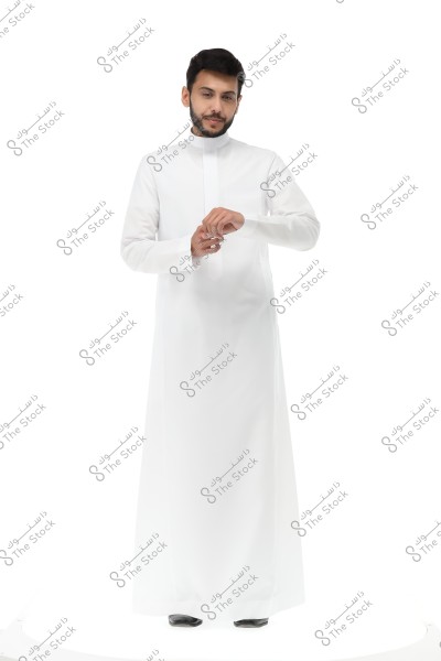 A photo of a man standing, wearing a white thobe, traditional attire indicative of Saudi or Gulf culture. The man appears to be adjusting his watch or cuffs, standing against a white background.