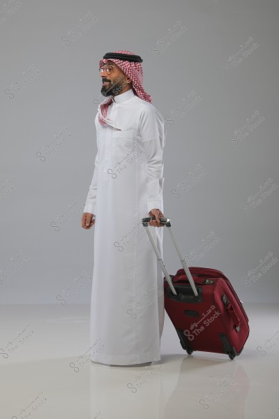 A man wearing a white thobe and a red checkered headscarf with a black agal, likely from Saudi Arabia, stands next to a red suitcase. The background is plain gray and the surface is slightly reflective.