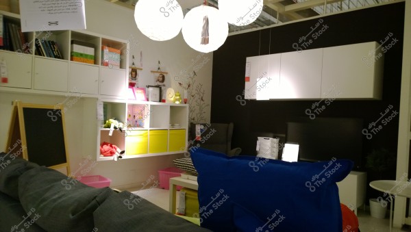 An image of an interior showroom displaying a modern room design. The scene shows a room decorated with vibrant colors, including a gray sofa with a blue cushion in the foreground, a white coffee table, and white storage units with colorful yellow and pink cubes on the back wall. There are also chairs, shelves with books, toys, and some hanging pictures. The lighting consists of three large round hanging lamps from the ceiling.