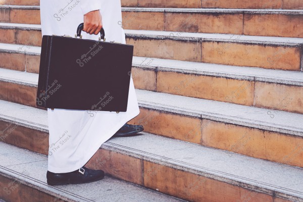 The image shows a person wearing traditional white clothing, likely from Saudi Arabia, walking up stairs while holding a black briefcase.