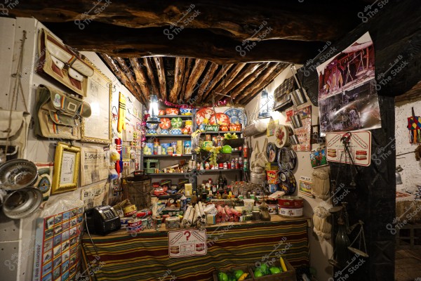 Image of a traditional store filled with vintage goods. The store has shelves stocked with various products like cans, jars, and packages. The walls are adorned with old photos and ads, along with bags and manual tools like scales. The ceiling is made of wood, and the space is lit by hanging lanterns.