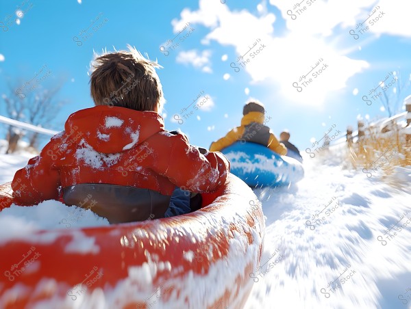 A child in a red jacket sits in a red snow tube, accompanied by other children in warm clothing, sledding down a snowy hill. The bright sun shines down, and the sky is clear blue with scattered white clouds.