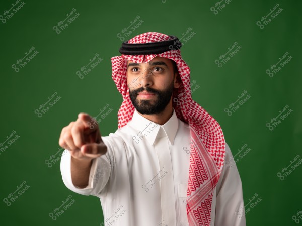 A portrait of a man wearing a white thobe and a red checkered headscarf with a black agal, pointing his finger towards the camera. The background is green, and the traditional clothing suggests he is from Saudi Arabia.