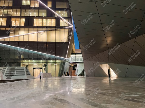 Image of a modern building with a unique architectural design, featuring a glass facade illuminated by night lights, with clear geometric details in its structure. The foreground floor has neutral, polished colors, emphasizing the geometric lines in the overall design of the building.