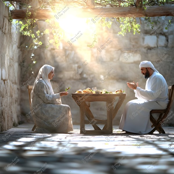 A man and a woman are sitting at a small wooden table with various dishes of food. The man, dressed in traditional white clothing and a turban, faces the woman who is wearing a hijab and a patterned dress. The setting is enclosed by stone walls, with sunlight filtering through the leaves above, casting beautiful shadows on the ground.