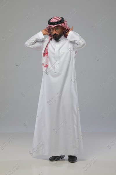 صورة لرجل يقف أمام خلفية رمادية, يرتدي ثوباً أبيض مع شماغ أحمر وأبيض على رأسه ومثبت بعقال أسود. الرجل يرتدي نظارات ولديه لحية, ويقوم بتعديل الشماغ بيديه.