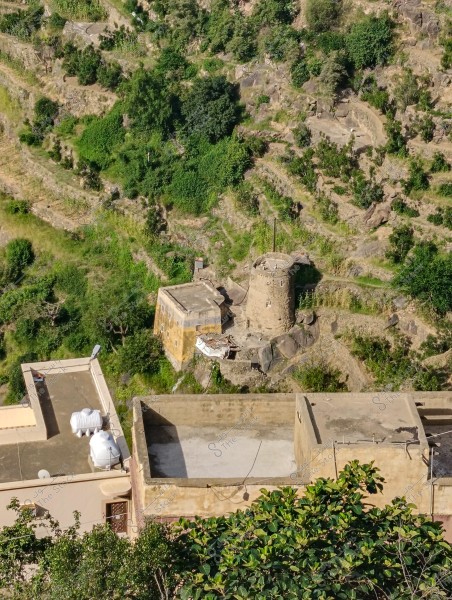 An image of a natural landscape featuring terraced green hills in a rural setting. The picture includes traditional stone and mud houses, some with water tanks on the roofs. In the center, there is an old, circular tower surrounded by trees and lush vegetation.
