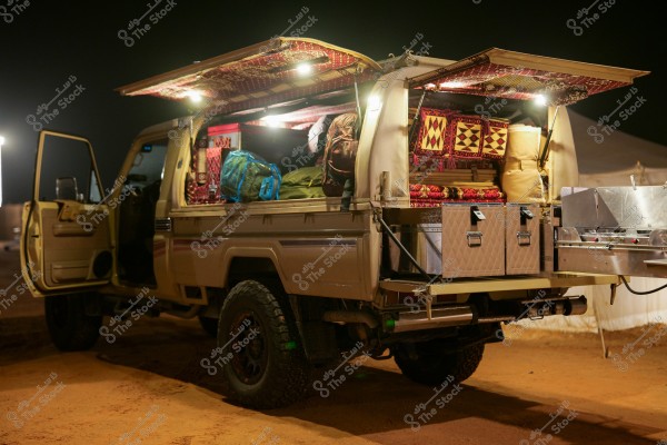 The image shows an off-road desert camping vehicle parked on a sandy ground under dim lighting. The rear of the vehicle is open, displaying meticulously organized camping equipment, including silver storage boxes, cushions with traditional Arabic patterns, backpacks, and various other bags. The side doors of the vehicle are also open, reflecting a serene desert environment prepared for nighttime adventures.