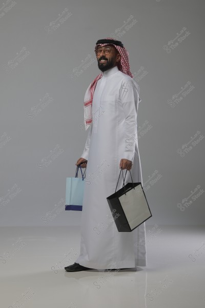 Image of a man wearing traditional Gulf attire, including a thobe, ghutra, and agal, standing and smiling while holding shopping bags in a studio environment with a gray background.