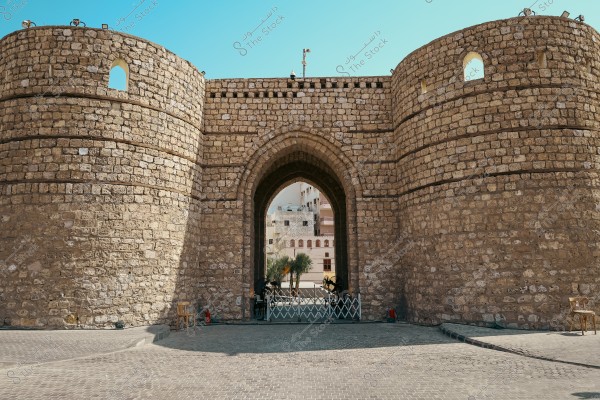 An image of a grand historical gate made of brown stones with a large central arch. The gate resembles traditional architectural style from the Arabian Peninsula. Palm trees and modern buildings are visible in the background through the central opening of the gate. The sky is clear blue, adding brightness and vibrancy to the scene.