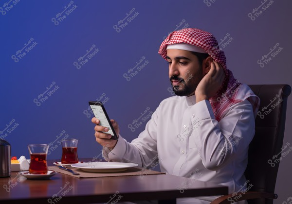 An image of a man wearing traditional Saudi attire, sitting at a table and holding a mobile phone. He is dressed in a white thobe and a red and white headscarf. On the table, there are two glasses of tea, an empty plate, and butter, under dim lighting with a blue background.