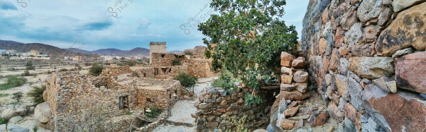 The image shows an old stone-built village in a mountainous area. The village consists of several adjoining stone buildings and narrow streets surrounded by stone walls. A large tree with dense foliage is visible on the right side, while a modern village can be seen in the background with mountain hills under a cloudy sky.