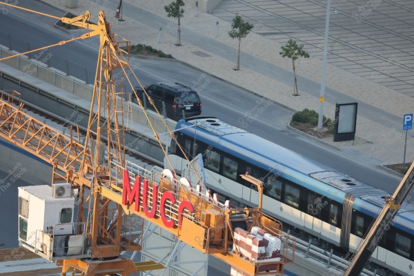 The image depicts an urban scene with a modern train running on an elevated track beside a roadway. In the foreground, there is a large construction crane with a prominent sign reading \"MUCC.\" The road is lined with small trees and geometrically designed pavements. A black SUV is driving on the road next to the train.