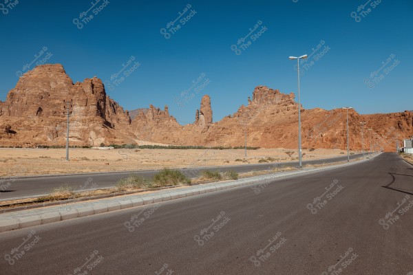 A wide paved road stretches through a rocky terrain and brown-colored mountains in a desert area. The mountains feature unique rock formations under a clear blue sky. There are streetlights along the side of the road.