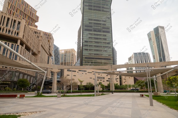 The image shows a view of a modern architectural complex featuring high-rise buildings with glass facades and neutral-colored patterns. The foreground is tiled and landscaped with some trees and green plants. Shadows extend across the area, created by the unique design of columns and overhead structures. The weather is cloudy, adding a calm appearance to the scene.